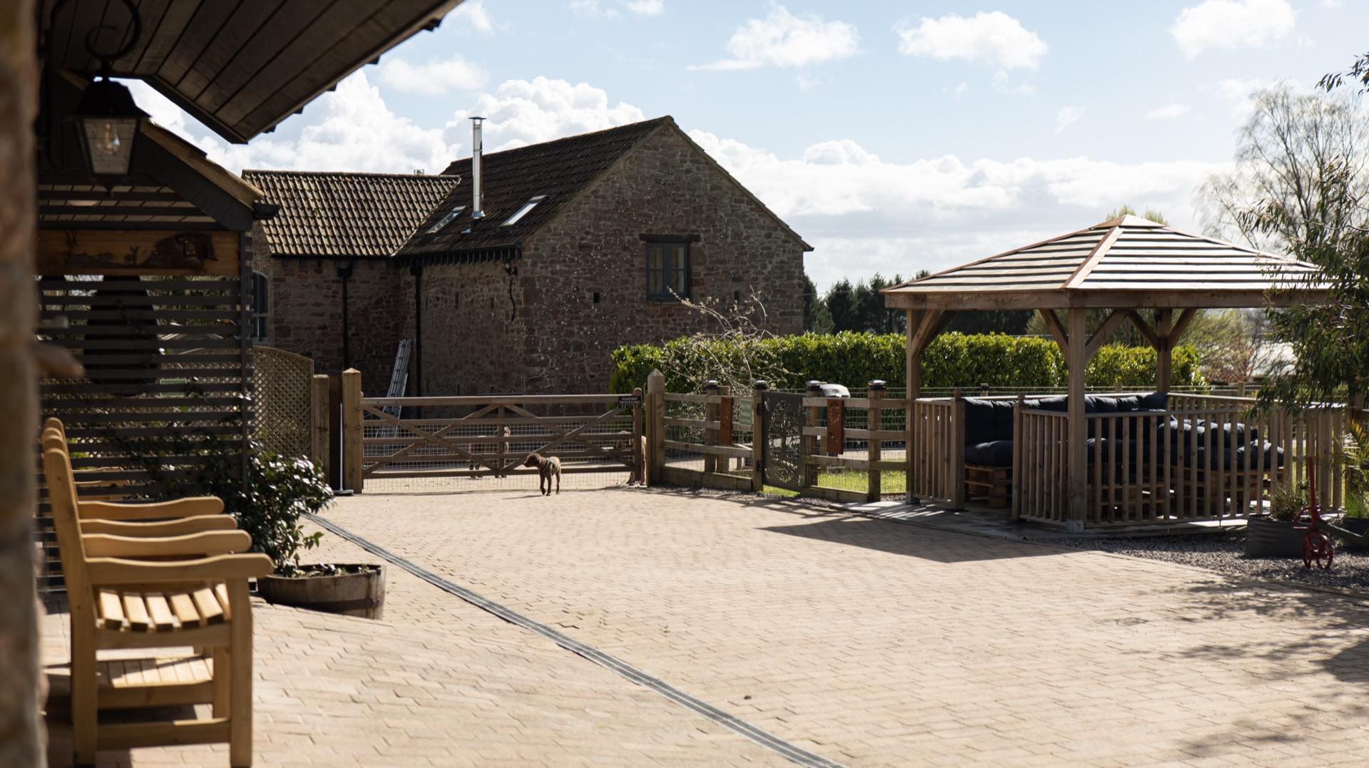 Paved courtyard with wooden benches, a five-bar gate, timber gazebo and a stone barn behind, with a small dog on the paving