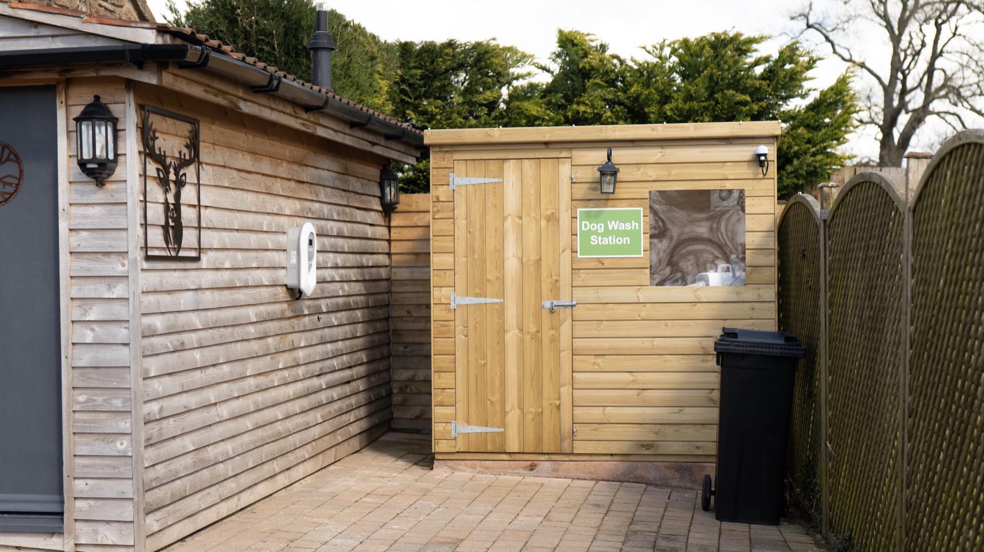 Small timber outbuilding with a green Dog Wash Station sign beside a timber-clad building on paving