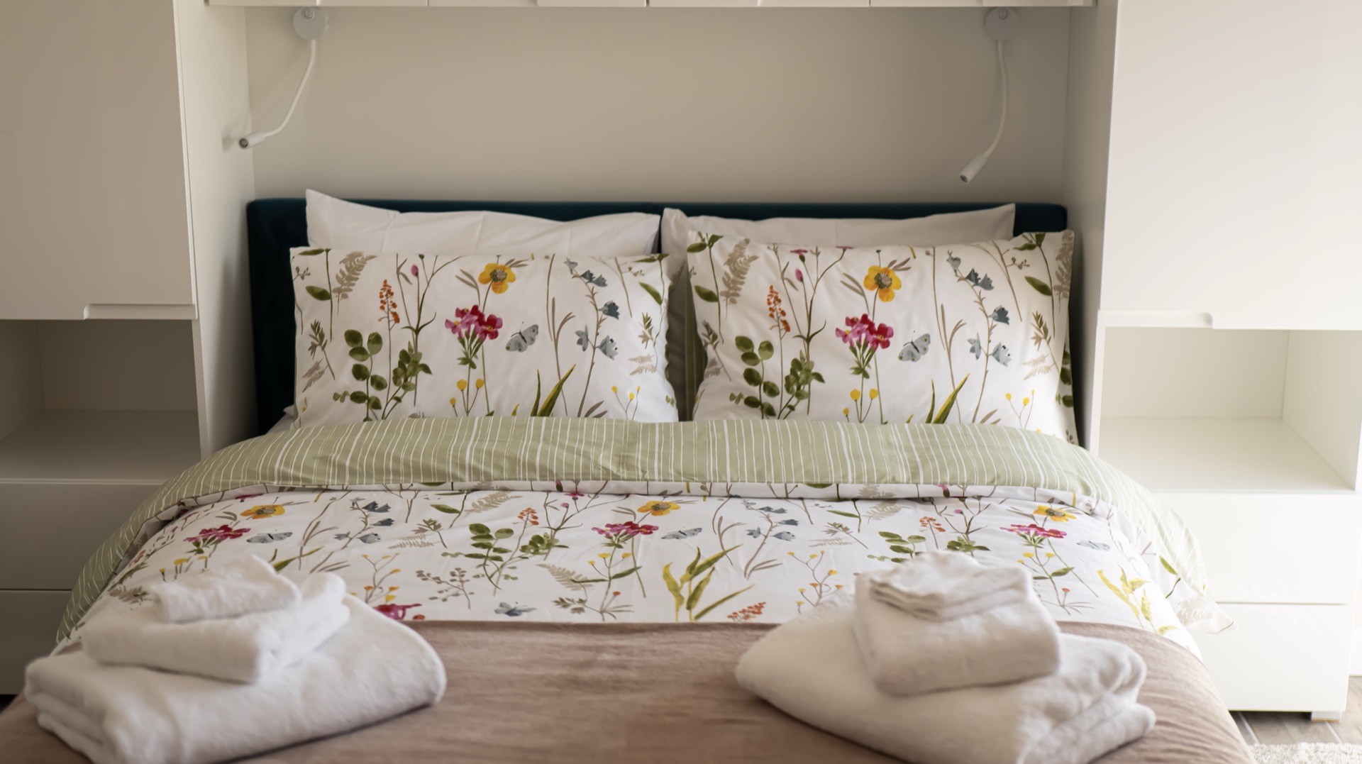 Elm double bed with wildflower-print bedding, teal headboard, folded white towels and white overhead cabinets either side