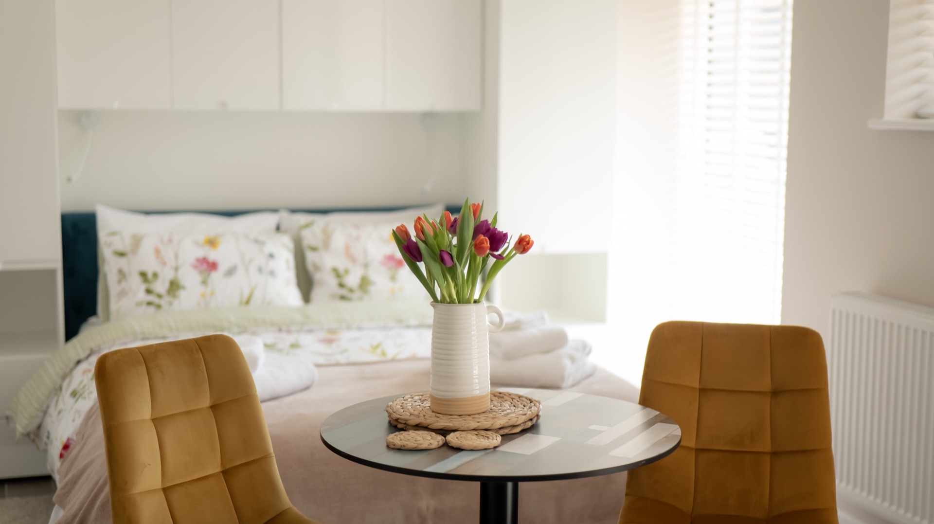 Round bistro table with a jug of tulips and two mustard chairs in Elm, double bed softly focused behind