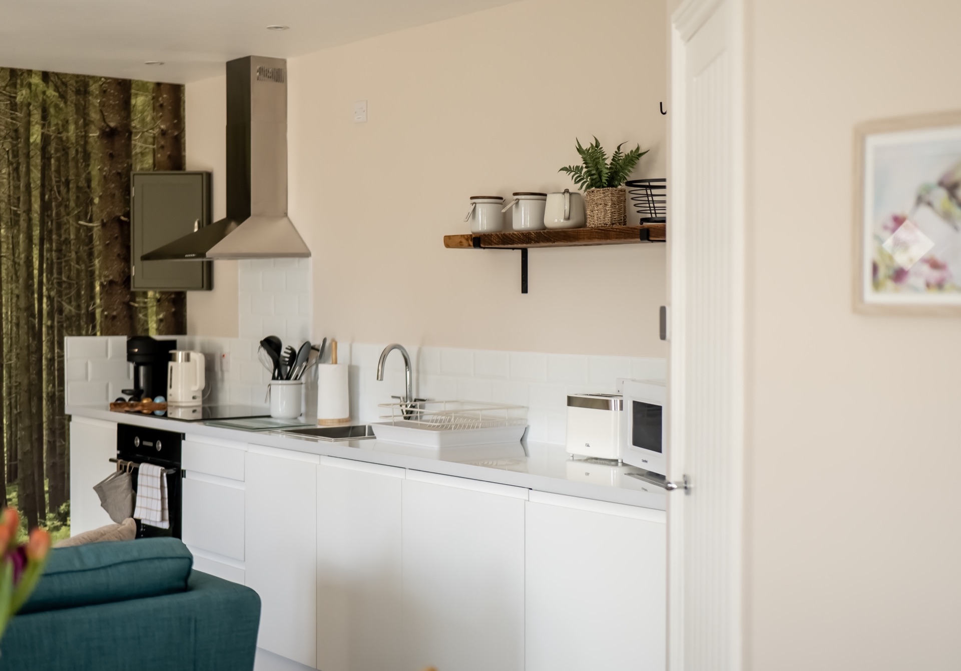Elm kitchen with white handleless units, stainless extractor hood, open wooden shelf with ceramics and a forest mural on the end wall