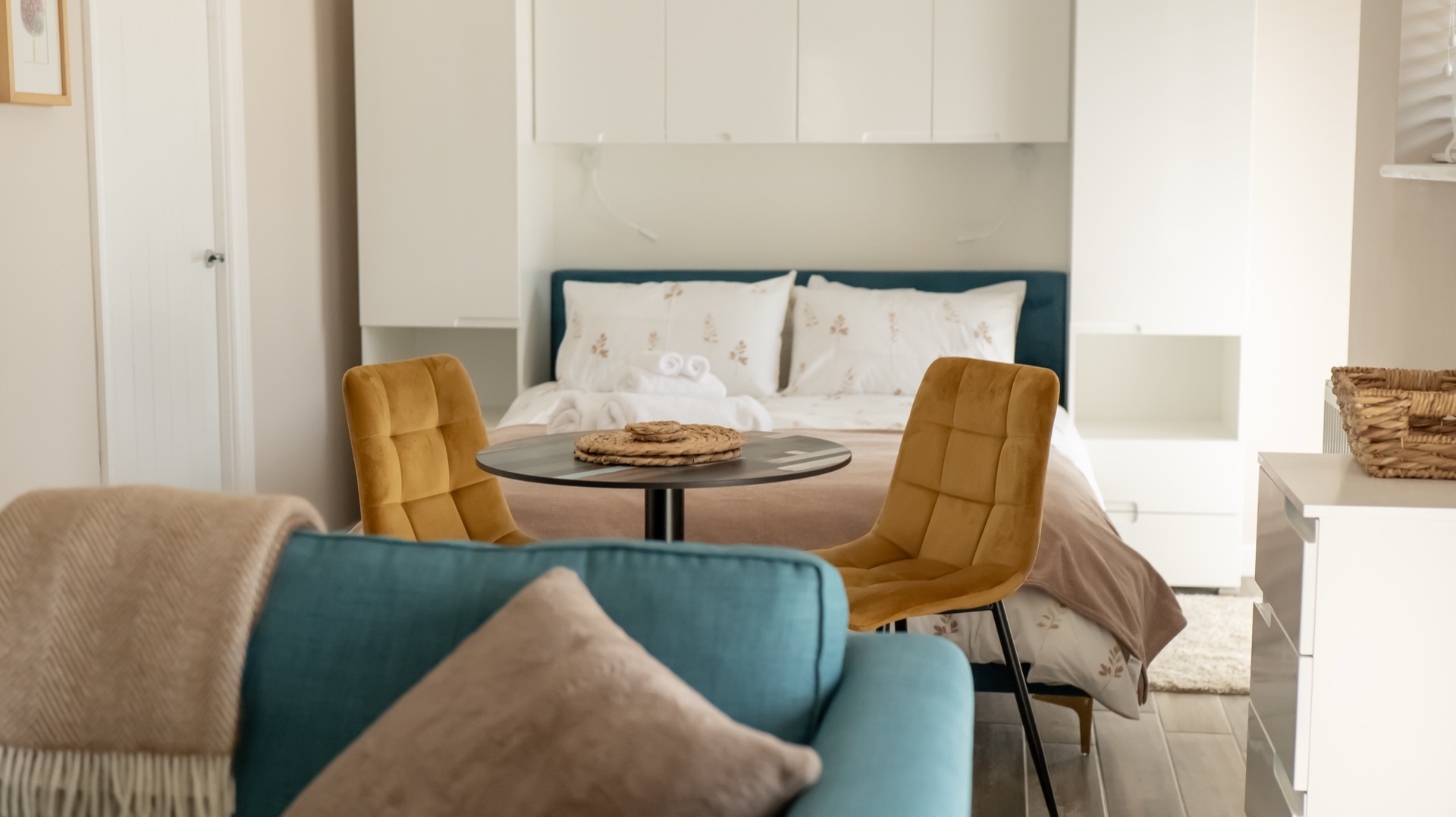 Elm studio interior with teal sofa and herringbone throw in the foreground, dining table, mustard chairs and double bed behind