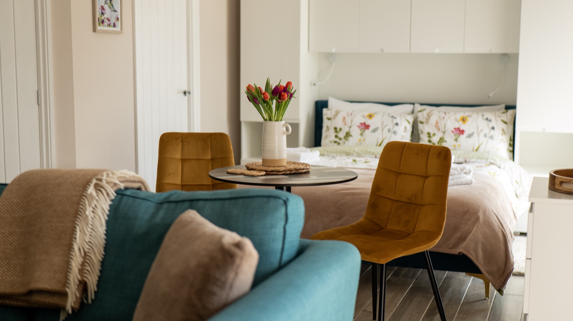Elm open-plan living space with teal sofa, round bistro table with tulips, two mustard velvet chairs and the double bed beyond