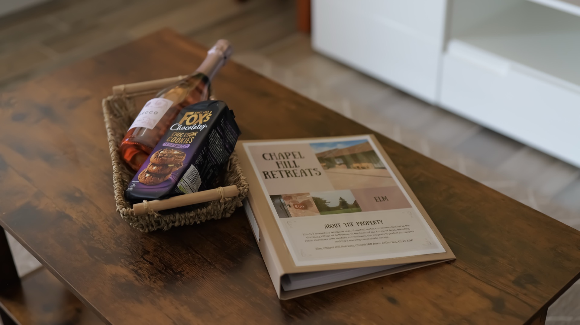 Welcome basket with a bottle of prosecco and cookies beside the Elm guest information folder on a wooden table