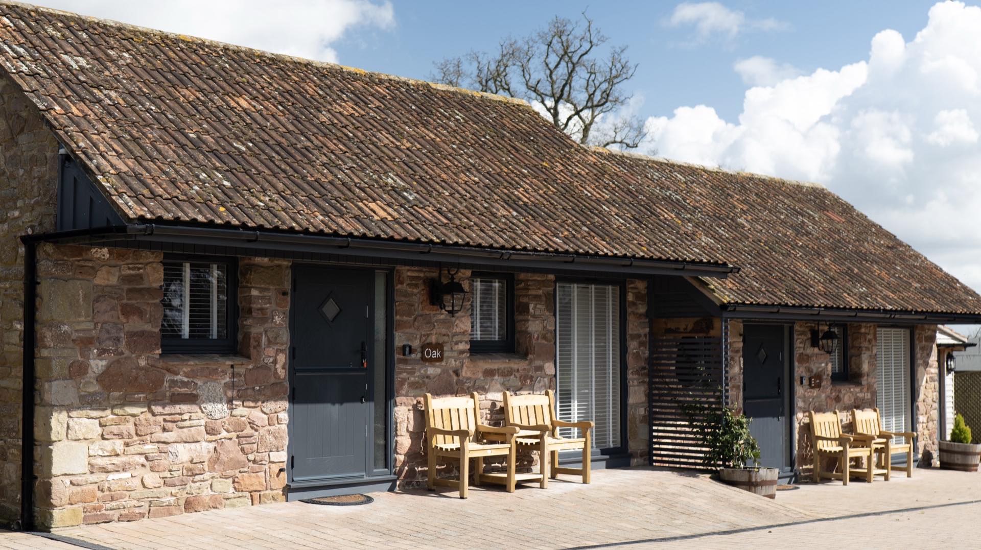 Stone barn frontage with a grey stable-style door, wooden armchairs and slatted timber screen on a paved terrace