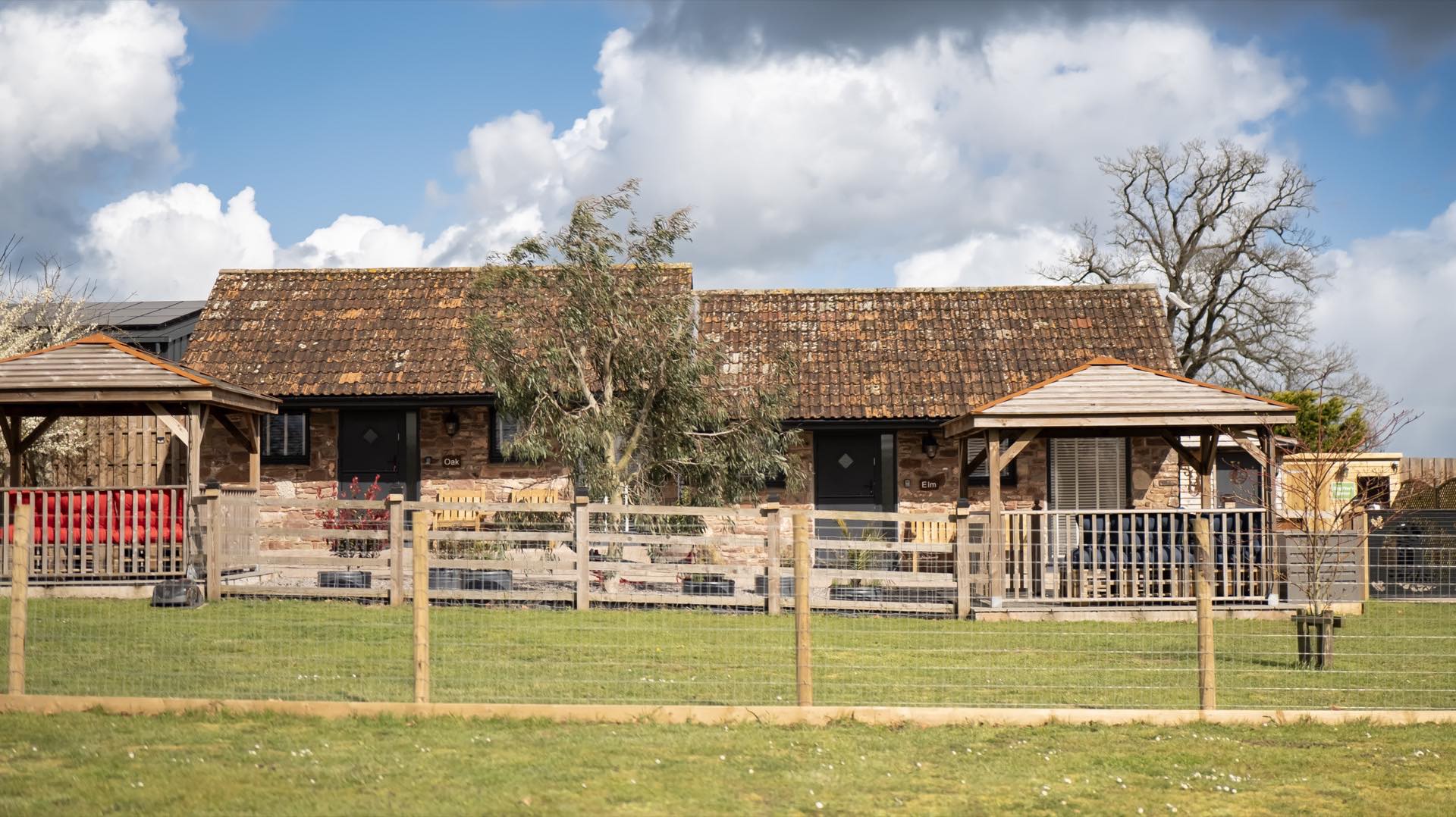 Single-storey stone barn conversion with tiled roof, covered seating gazebos and a fenced lawn in front