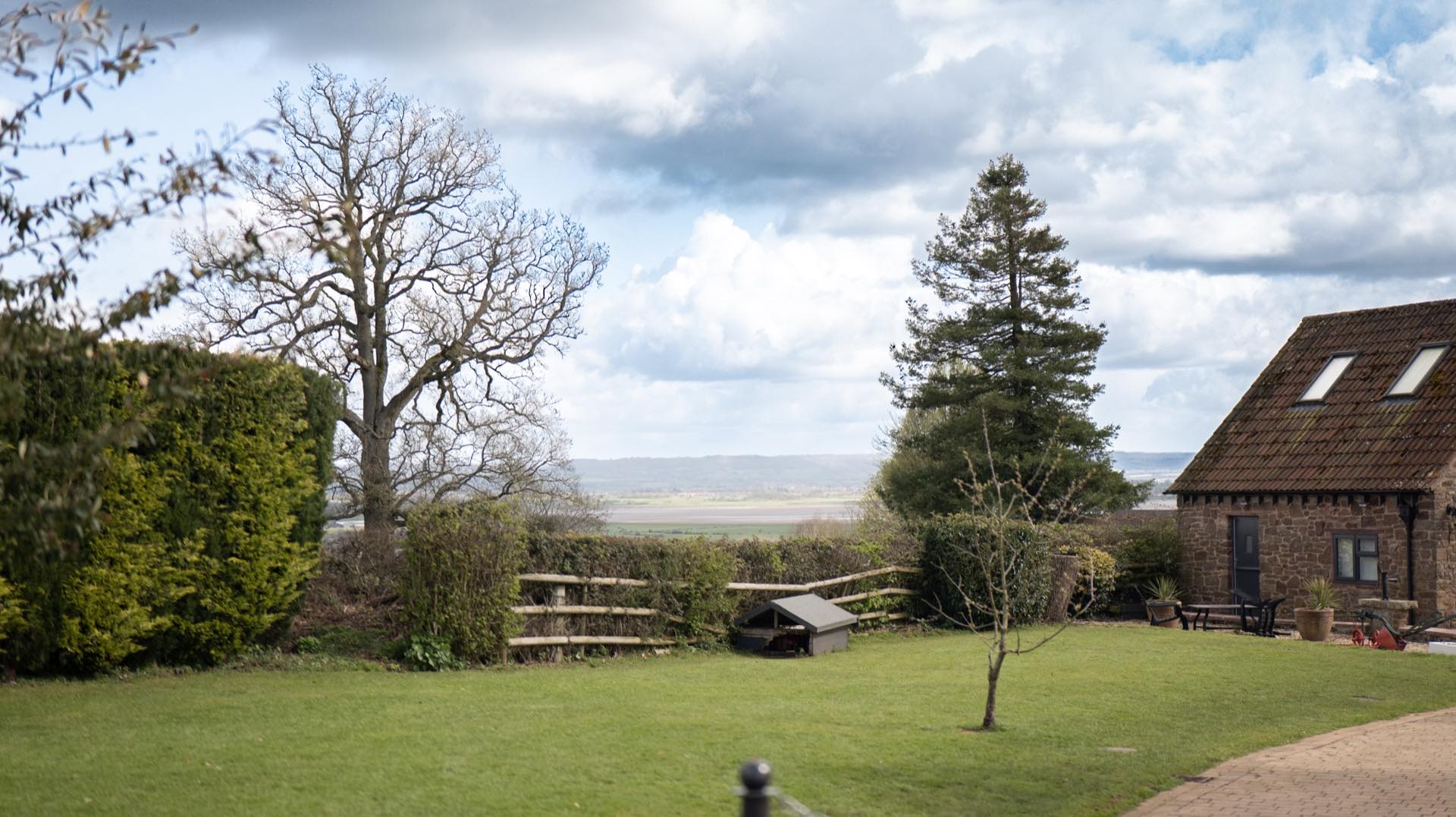Lawned garden with clipped hedges, post-and-rail fencing and a stone outbuilding, with open countryside beyond