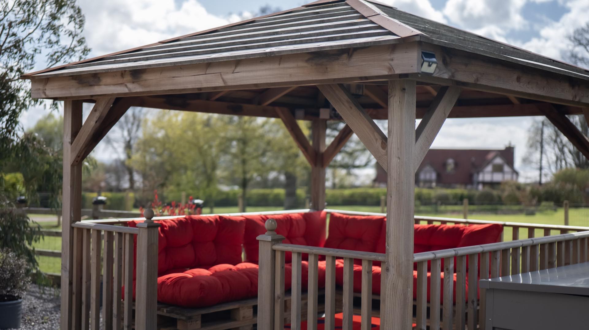 Timber gazebo with slatted roof and built-in seating with deep red cushions, looking out over fenced grass and trees