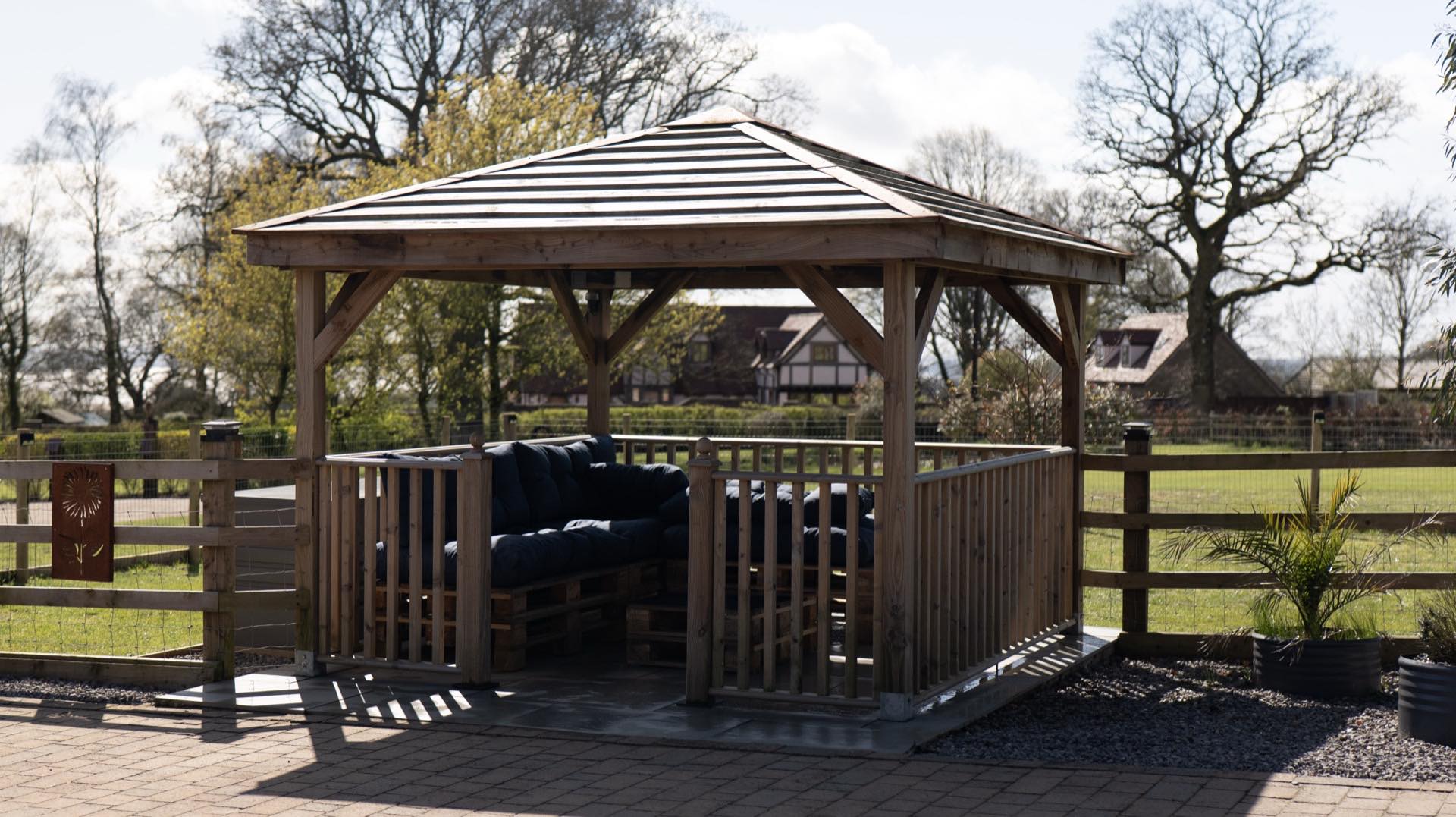 Timber gazebo with slatted roof and built-in seating with navy cushions on a paved area, fenced grass and trees behind