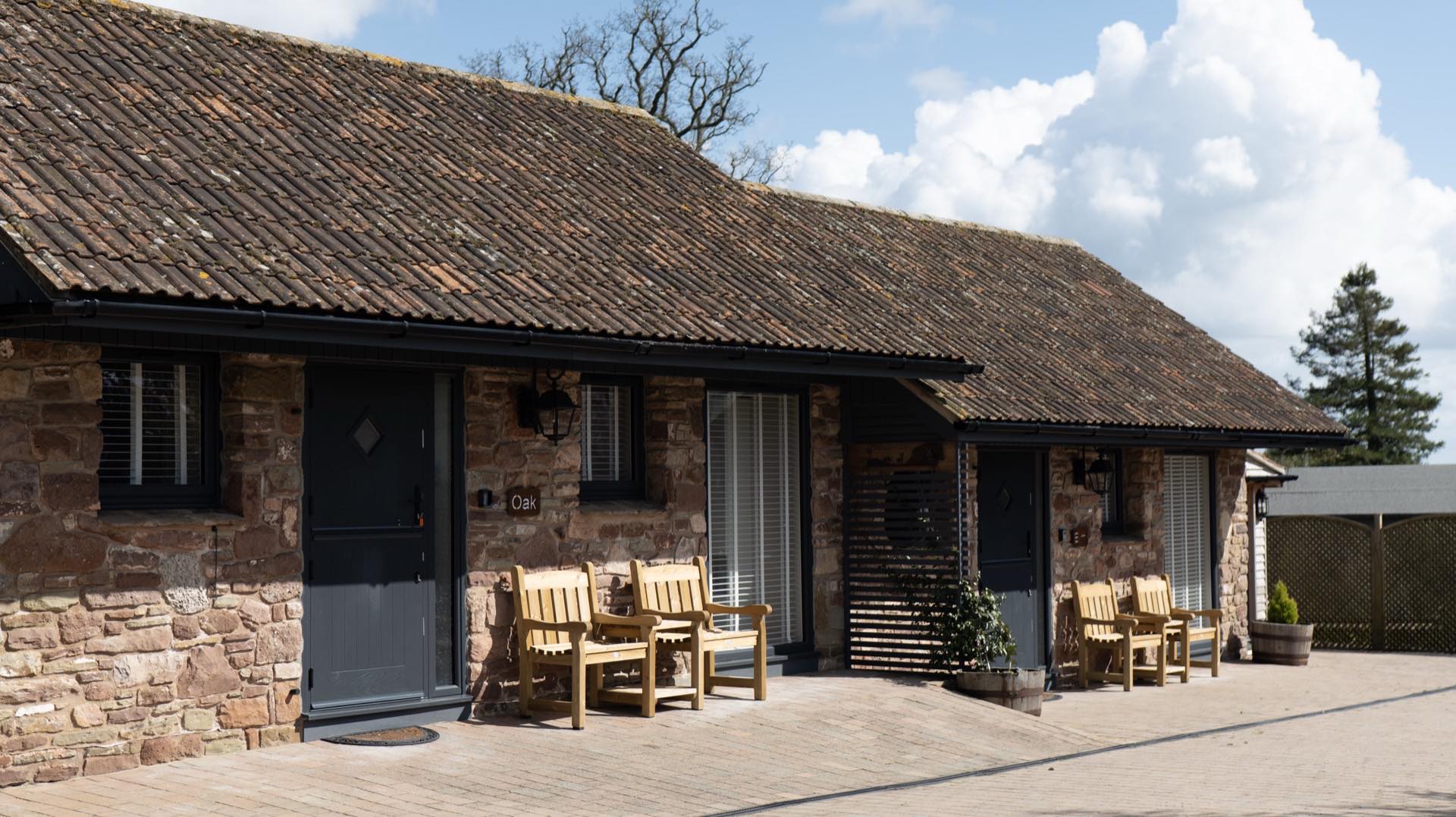 Front elevation of a stone barn conversion with dark doors and pairs of wooden armchairs on a block-paved terrace
