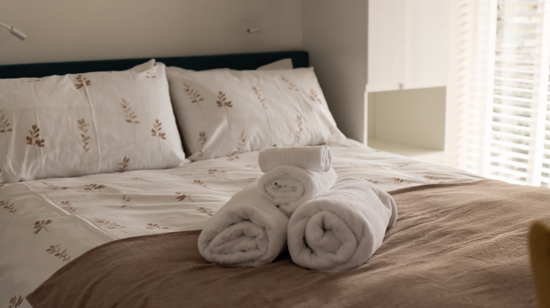 Rolled white towels on the Oak bed with leaf-print bedding and soft light through the window blinds