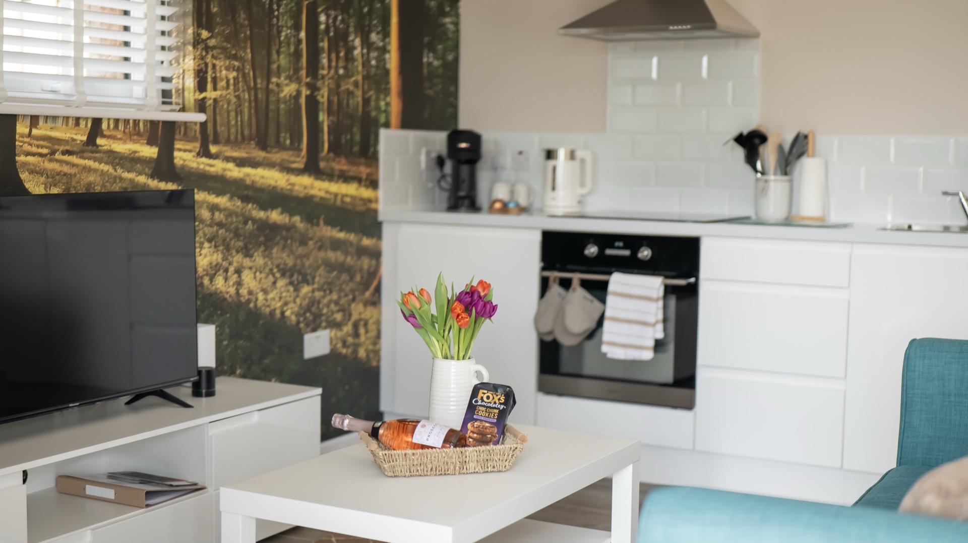 Oak living area with television, white kitchen units, woodland photo mural wall and a welcome basket on the coffee table