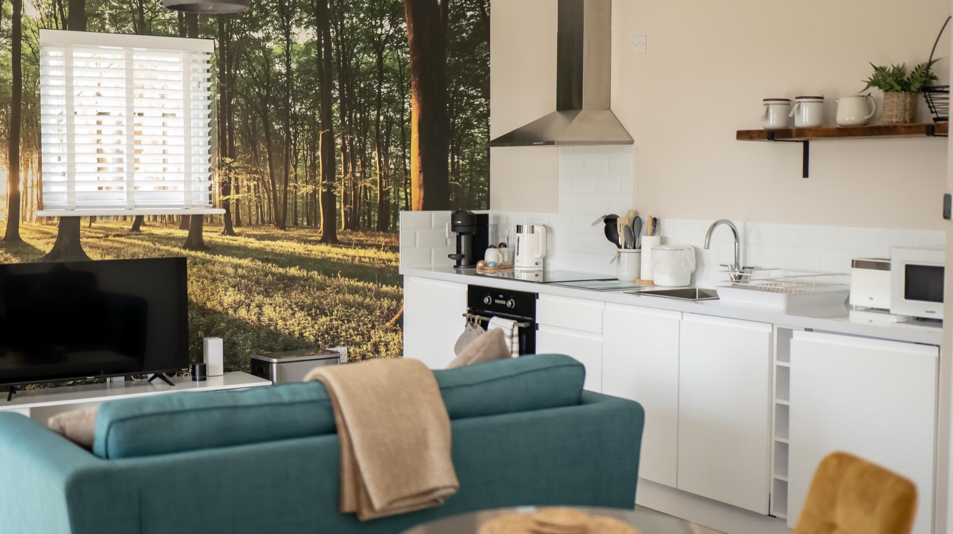 Wide view of the Oak open-plan living space with woodland mural wall, television, white kitchen and teal sofa