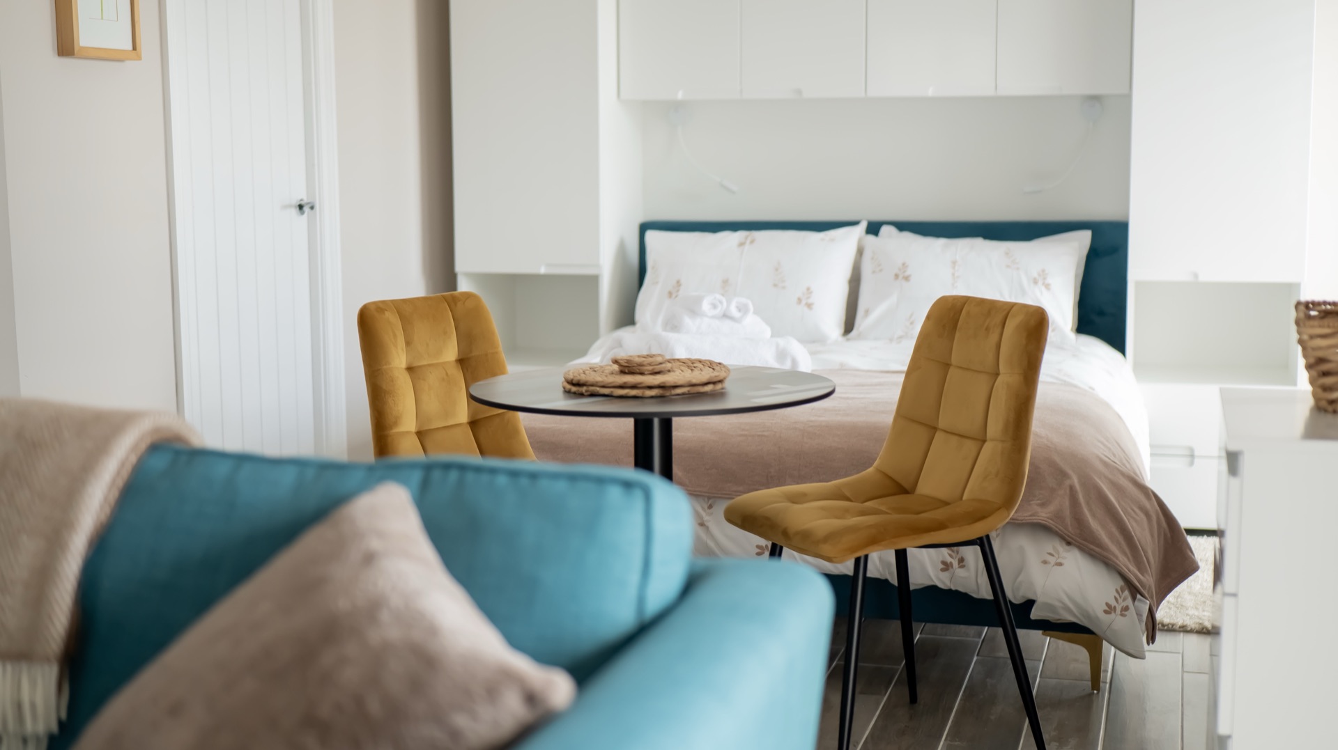 Oak studio with double bed, round dining table with two mustard velvet chairs, and teal sofa in the foreground