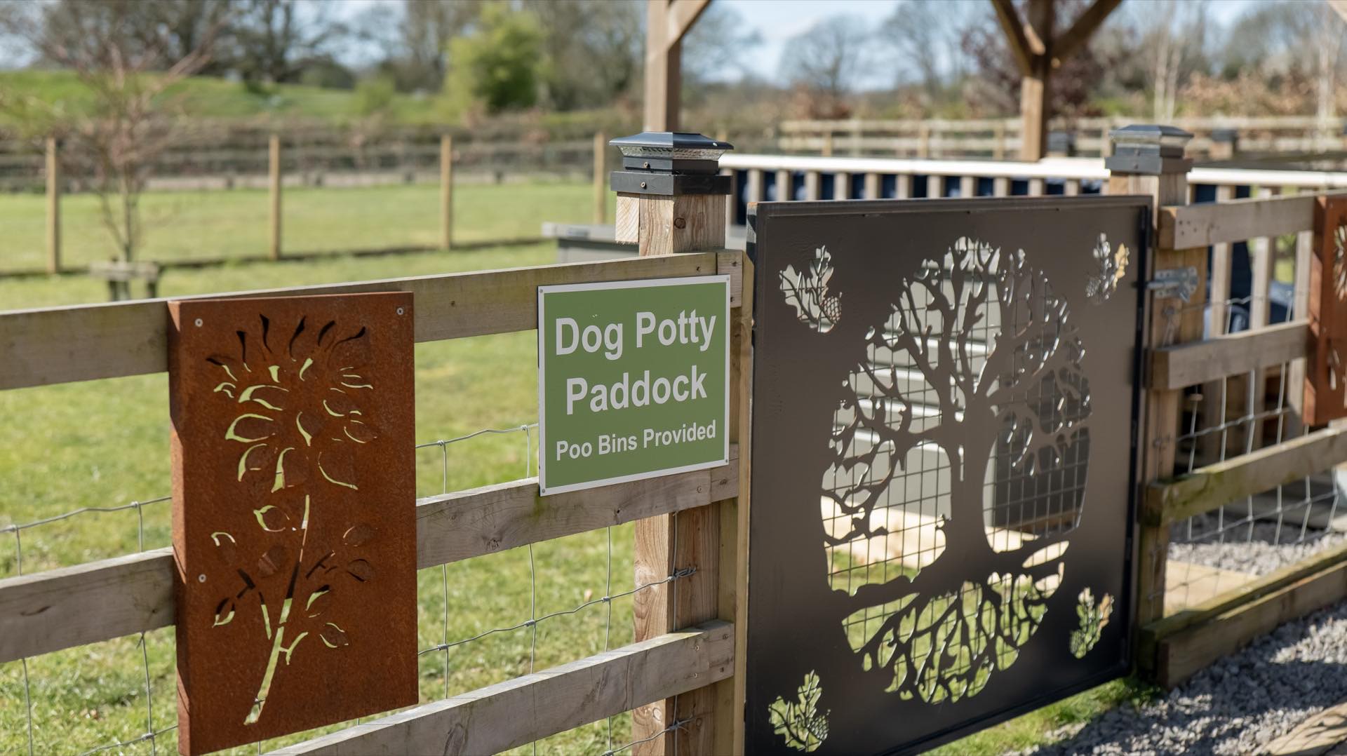 Paddock sign and floral metal panel on post-and-rail fencing with a decorative gate and grass paddock beyond