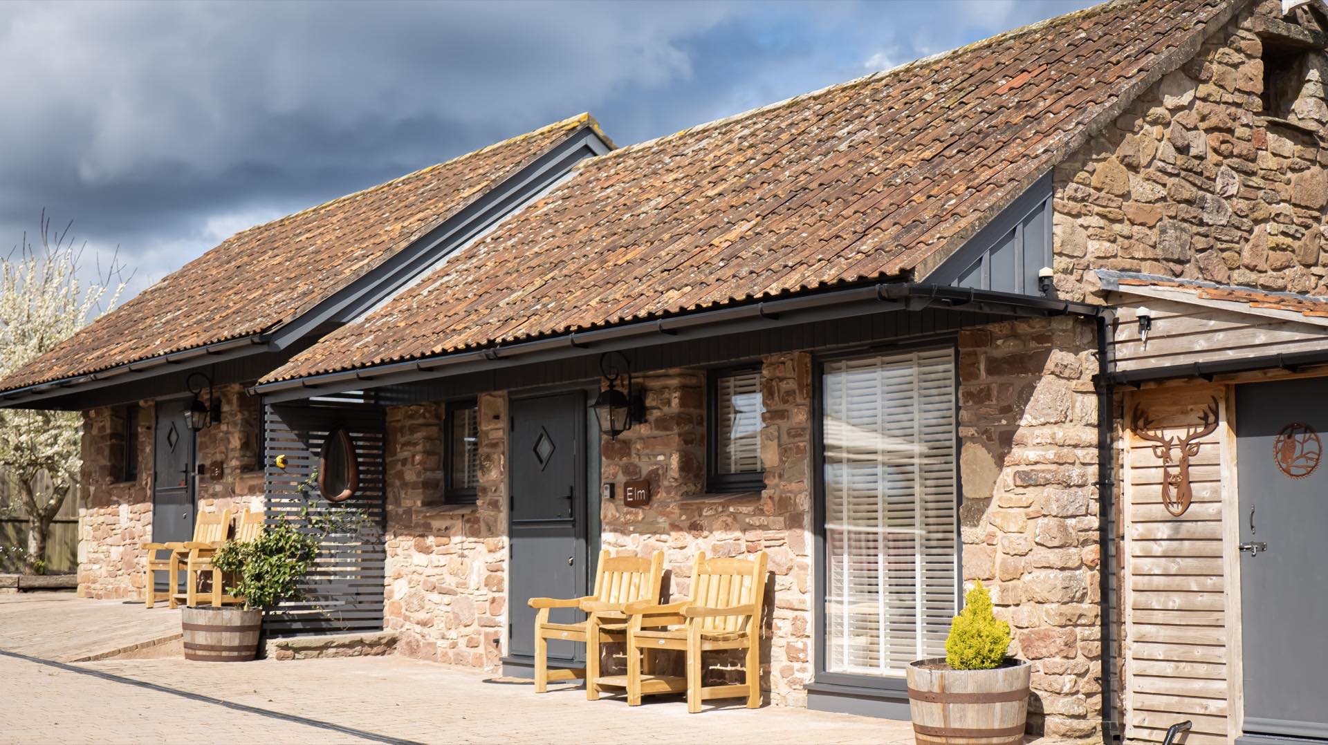 Stone barn frontage with grey stable-style doors, lantern wall lights, wooden armchairs and barrel planters on a paved terrace